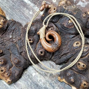 May include: A wooden pendant in the shape of a fish hook, with a carved design, hangs from a brown cord necklace. The pendant is brown and has a natural wood finish. The necklace is made of a braided cord.
