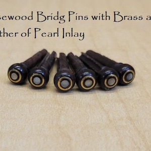 May include: Rosewood bridge pins with brass and mother of pearl inlay. The image shows six dark brown bridge pins with a brass ring and a white mother of pearl inlay. The text in the image reads "Rosewood Bridge Pins with Brass and Mother of Pearl Inlay."