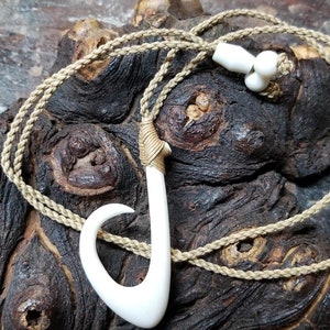 May include: A white bone fish hook pendant on a brown braided cord necklace. The necklace has white beads at the top.