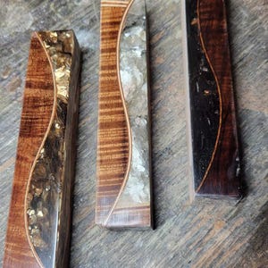 May include: Three wooden blocks with a wavy design and embedded glitter. The blocks are different colors: brown with gold glitter, brown with white glitter, and black with copper glitter.