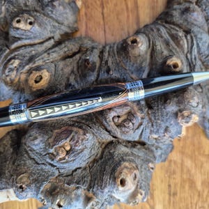 May include: A ballpoint pen featuring a dark wood grain barrel with a polished black finish. The pen includes a silver-toned metal clip decorated with a series of white triangular inlays and silver-toned bands with a textured crosshatch pattern.