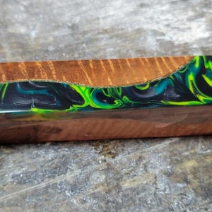 May include: A wooden block with a green, blue, and black resin inlay. The resin has a swirling pattern. The wood is a light brown color.