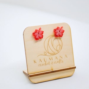 May include: A pair of red hibiscus flower earrings displayed on a wooden stand with the text "KAUMANA crochet & crafts".