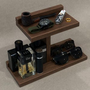 May include: A two-tiered dark wood organizer stand holding a tobacco pipe, a wristwatch with a green strap, a multi-tool, a gold ring, two black perfume bottles labeled Dior Homme and Chanel, and a pair of black sunglasses on the lower shelf.