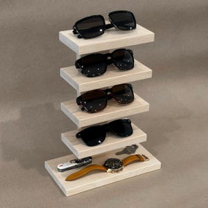 May include: A light-colored wooden display stand with four horizontal shelves holding four pairs of dark-tinted sunglasses. The base shelf holds a small multi-tool, a metal key, and a wristwatch with a tan leather strap.