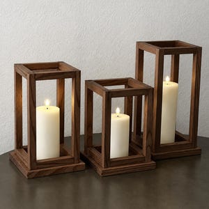 May include: Three rectangular wooden candle lanterns of varying heights, each featuring a dark brown stained finish and an open-frame design. Inside each lantern sits a single, smooth, cream-colored pillar candle with a lit flame at the top.