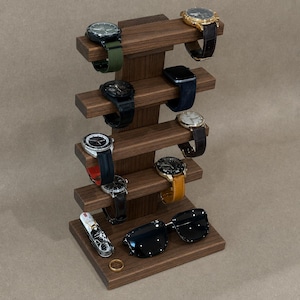 May include: A dark brown wooden watch stand with a vertical central pillar and six horizontal arms holding various wristwatches with different straps. The base features a multi-tool, a pair of black sunglasses, and a small gold-colored ring.