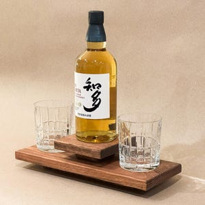May include: A bottle of amber-colored liquor with a white label and Japanese characters, accompanied by two clear, patterned glasses. The bottle and glasses rest on a tiered, dark wooden serving tray.