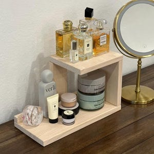 May include: A light-colored wooden tiered shelf holding various skincare and fragrance bottles, including a grey Verli bottle, a Jo Malone perfume, a Coco Chanel bottle, and two Salt & Stone jars, positioned next to a gold-rimmed vanity mirror.