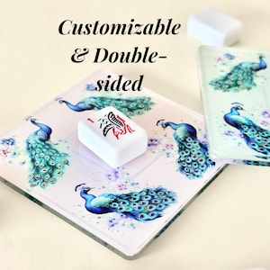 Könnte beinhalten: Anpassbare, doppelseitige quadratische Untersetzer mit Pfauendesigns in Blau- und Türkistönen. Auf einem Untersetzer liegt ein weißer Stein mit roten und schwarzen Symbolen. Der Text "Customizable & Double-sided" ist im Bild enthalten.
