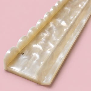 May include: A long, rectangular display stand crafted from off-white, pearlescent acrylic material. The stand features a scalloped edge along one side and a flat, recessed channel running the length of the piece, with a small, circular metal pin.