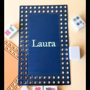 Pu&ograve; includere: Un sottobicchiere rettangolare blu navy con un bordo intrecciato e il nome "Laura" in bianco. Diverse tessere di Mahjong sono sparse intorno al sottobicchiere, con vari simboli e numeri.
