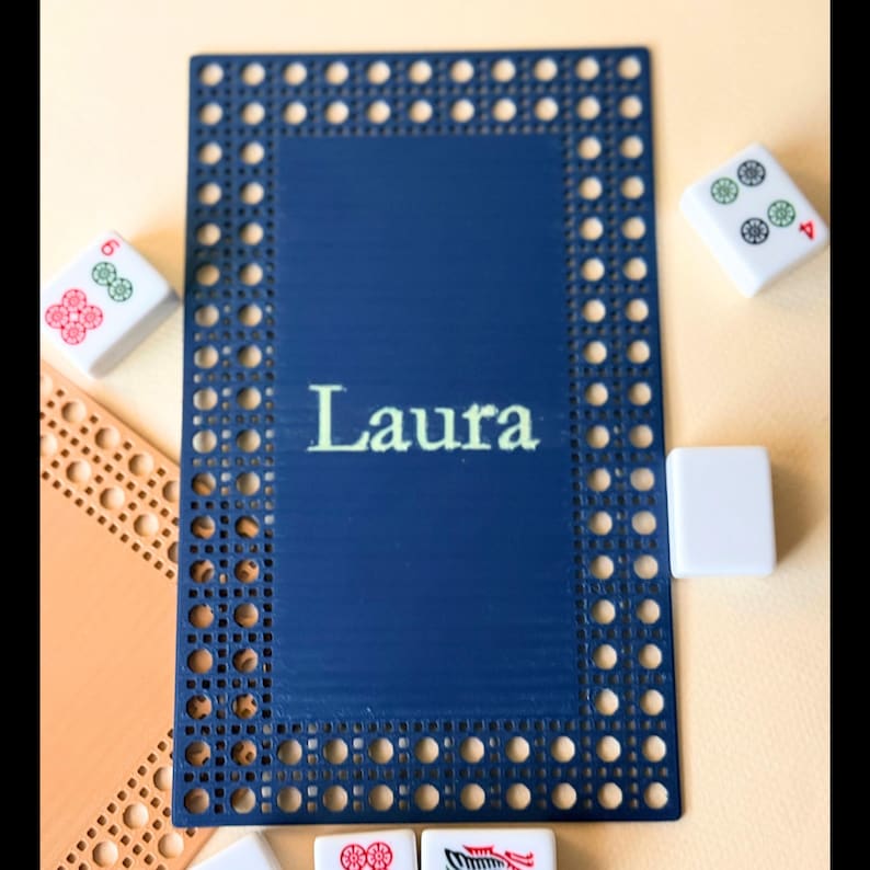 Pu&ograve; includere: Un sottobicchiere rettangolare blu navy con un bordo intrecciato e il nome "Laura" in bianco. Diverse tessere di Mahjong sono sparse intorno al sottobicchiere, con vari simboli e numeri.
