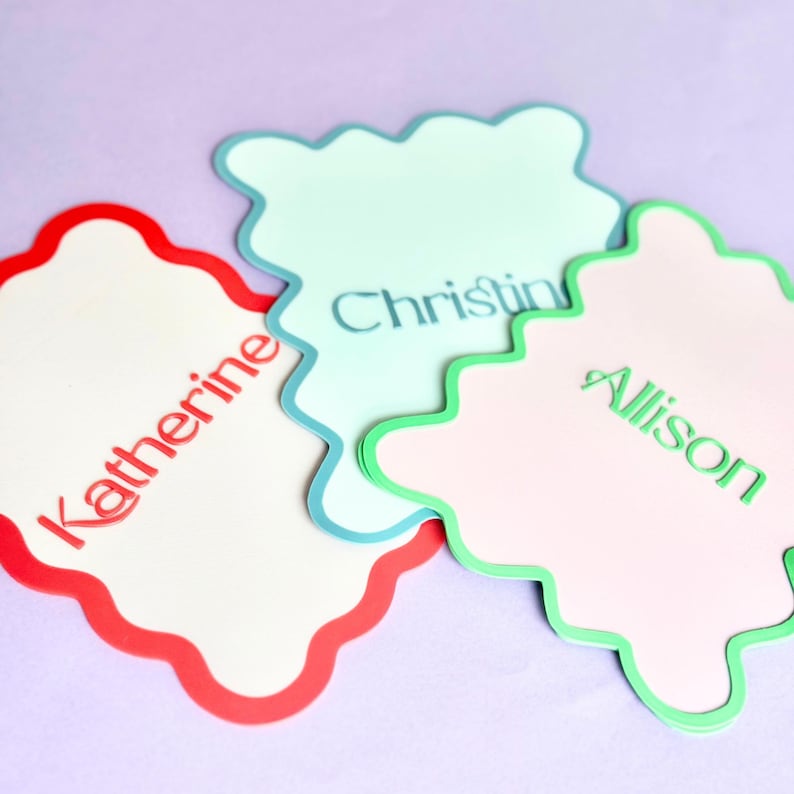 May include: Three scalloped-edge name tags in red, blue, and green. The name tags are white with the names "Katherine", "Christine", and "Allison" printed on them in red, blue, and green, respectively. The background is a light purple.