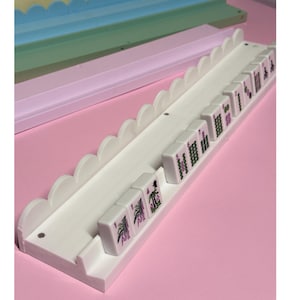May include: A white mahjong tile rack featuring a scalloped top edge and a horizontal channel for holding tiles. Several white mahjong tiles with green and pink printed symbols and numbers are arranged in a row along the rack's base.