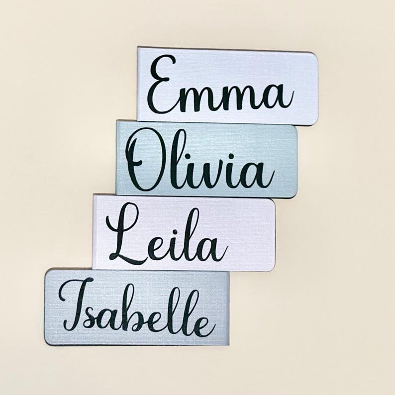May include: Four personalised name tags with the names Emma, Olivia, Leila, and Isabelle. The tags are made of a light grey material with a subtle texture and black lettering.