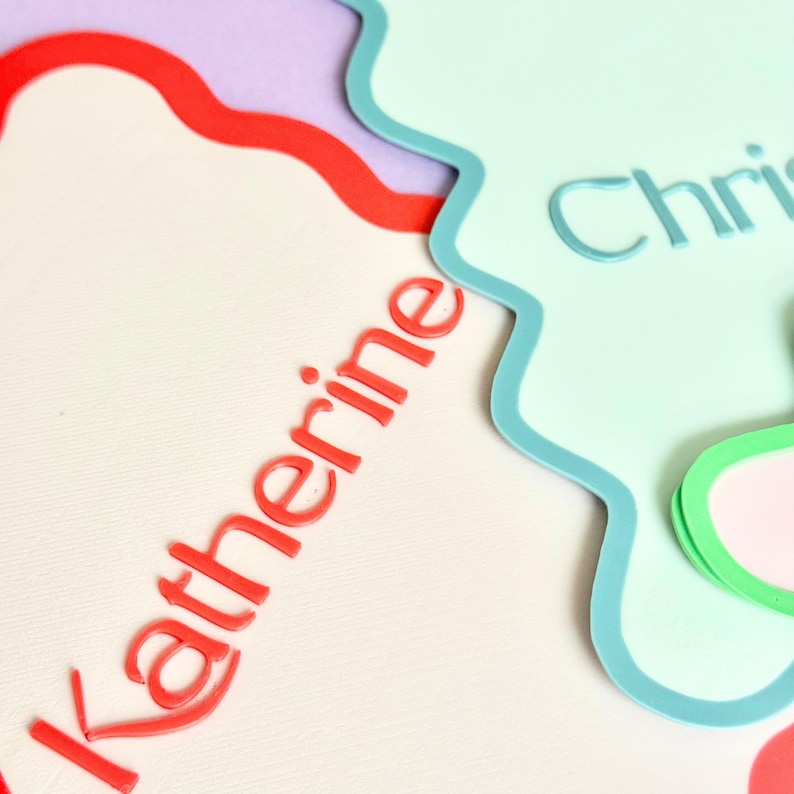 May include: Close-up of layered paper cutouts in pastel colors. The word "Katherine" is in red, and the word "Chris" is in blue. The cutouts have wavy edges and are arranged on a light cream background.