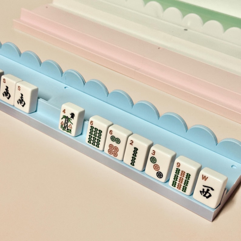 May include: A light blue mahjong tile rack with a scalloped top edge, holding a row of white mahjong tiles featuring various green, red, and black symbols, numbers, and letters. The rack is positioned in front of a pale pink tiered tray.