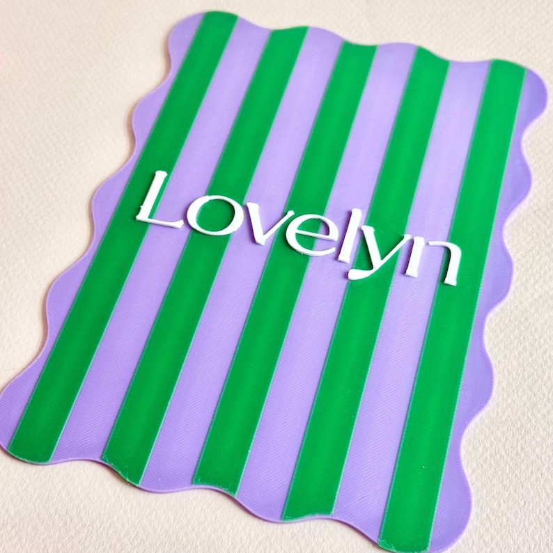 May include: A rectangular placemat with a wavy edge, featuring alternating green and lavender vertical stripes. The word "Lovelyn" is displayed in white, centered on the mat.