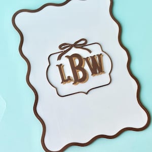 May include: A white, irregularly shaped wooden sign with a brown border. The sign features a smaller, decorative brown frame with the letters "LBW" in a brown and gold color scheme, topped with a bow.