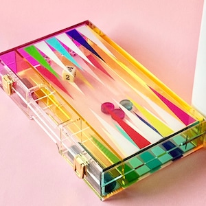 May include: A clear acrylic backgammon set with a vibrant rainbow design. The game board features colorful triangular patterns and includes dice and game pieces. The set has gold-colored clasps and a clear storage compartment.