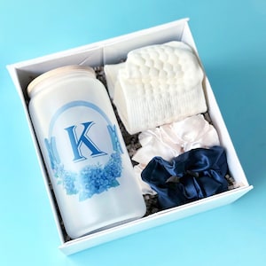 Puede incluir: Una caja de regalo blanca con un tarro de cristal esmerilado con un diseño floral azul y la letra "K". La caja también contiene calcetines blancos y dos coleteros de satén, uno blanco y otro azul marino. Sobre una superficie azul claro.