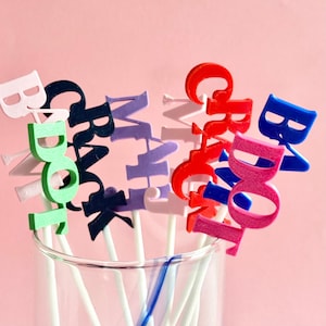 May include: A set of colorful, personalized drink stirrers with names printed on them. The stirrers are made of plastic and have white sticks. The names on the stirrers are "Brady", "Grace", "Whit", "Gracie", "Brady", and "Baby".