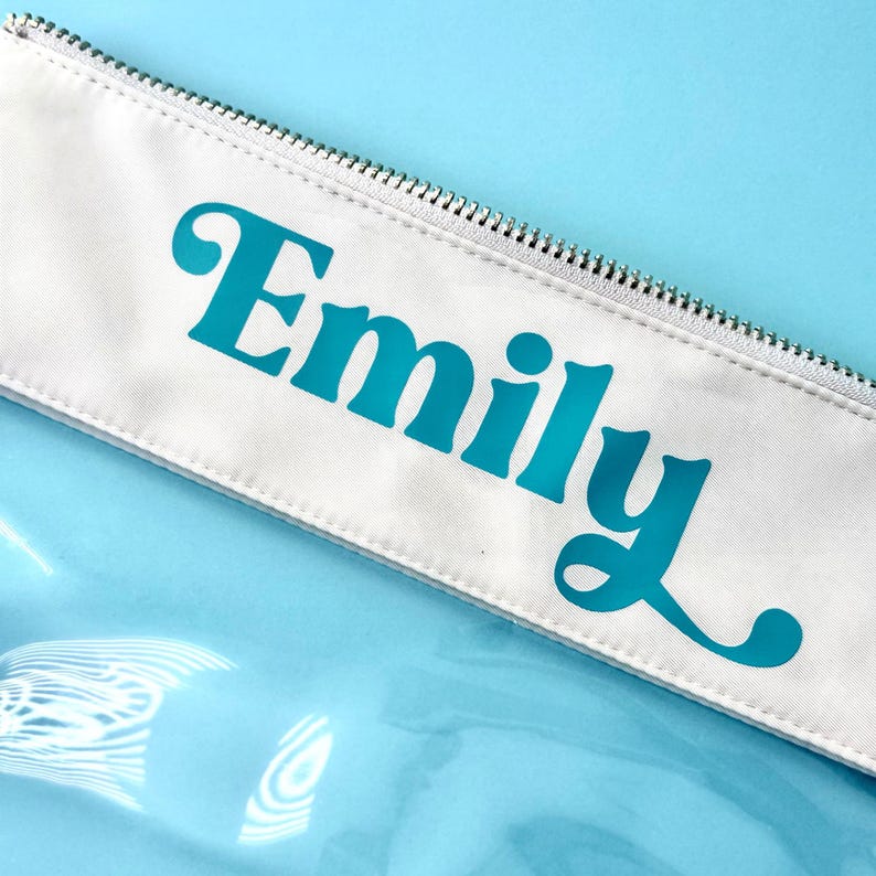 May include: White pencil case with a silver zip and the name "Emily" in teal script. The case is set against a light blue background. The design is minimalist and modern, suitable for stationery or cosmetics.