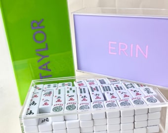 May include: A clear acrylic mahjong set with white tiles, displayed in a clear box. The set includes a green case with the name "TAYLOR" and a lavender case with the name "ERIN". The tiles are white with red, green, and black markings.