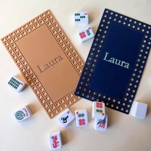 Pu&ograve; includere: Due tappetini da gioco rettangolari, uno beige e uno blu navy, con il nome "Laura" stampato sopra. Diverse tessere di Mahjong bianche con simboli rossi e verdi sono sparse intorno ai tappetini. I tappetini hanno un bordo decorativo.