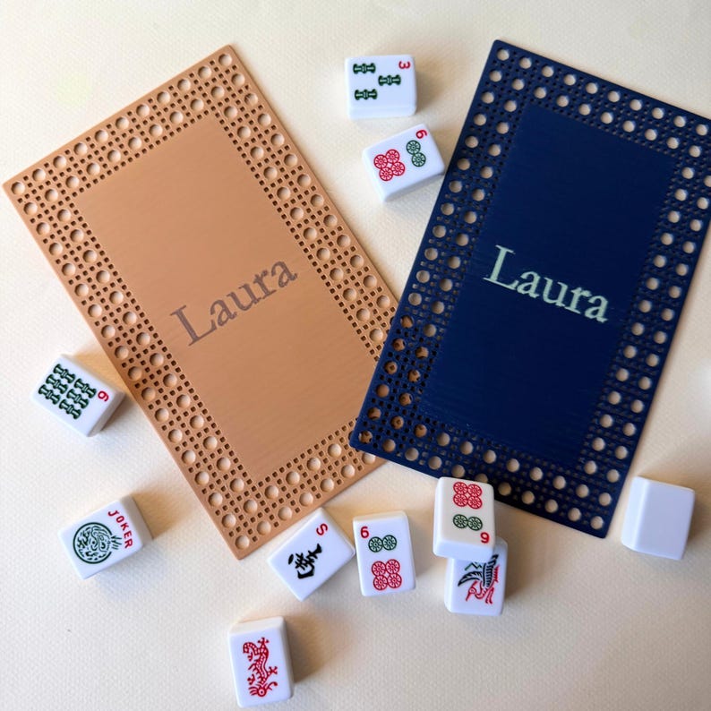 Pu&ograve; includere: Due tappetini da gioco rettangolari, uno beige e uno blu navy, con il nome "Laura" stampato sopra. Diverse tessere di Mahjong bianche con simboli rossi e verdi sono sparse intorno ai tappetini. I tappetini hanno un bordo decorativo.