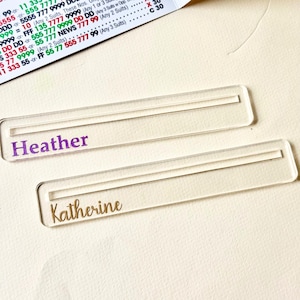 Puede incluir: Dos tarjeteros de nombre de acrílico transparente. Uno tiene el nombre "Heather" en morado y el otro "Katherine" en dorado. Los soportes son rectangulares con una ranura para una tarjeta. El fondo es de color beige claro.
