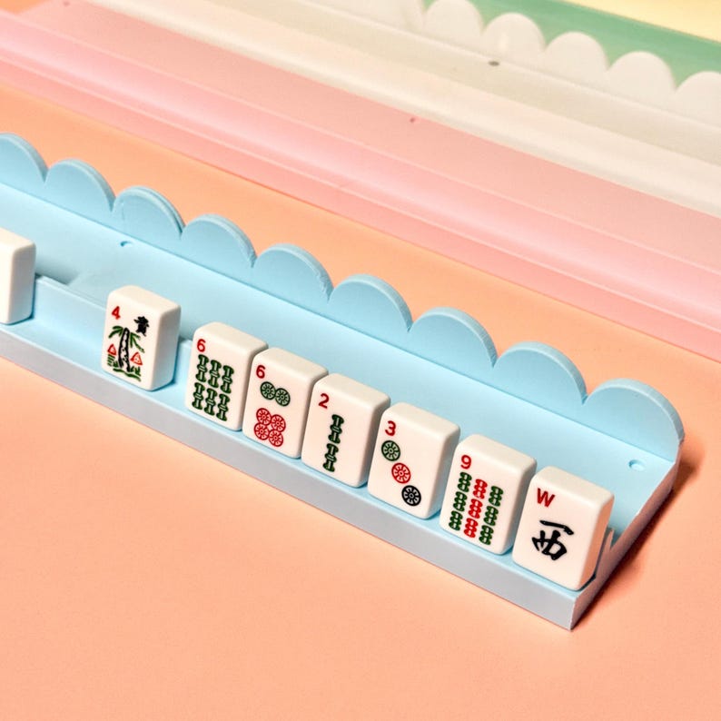May include: A light blue plastic Mahjong tile rack with a scalloped top edge, holding a row of white Mahjong tiles. The tiles display various green, red, and black symbols, including numbers, bamboo patterns, and the Chinese character for West.