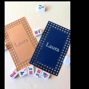 Pu&ograve; includere: Due porta tessere Mahjong personalizzati, uno color pesca e uno blu navy, con il nome "Laura" stampato sopra. I porta tessere hanno un bordo decorativo e sono circondati da tessere Mahjong.