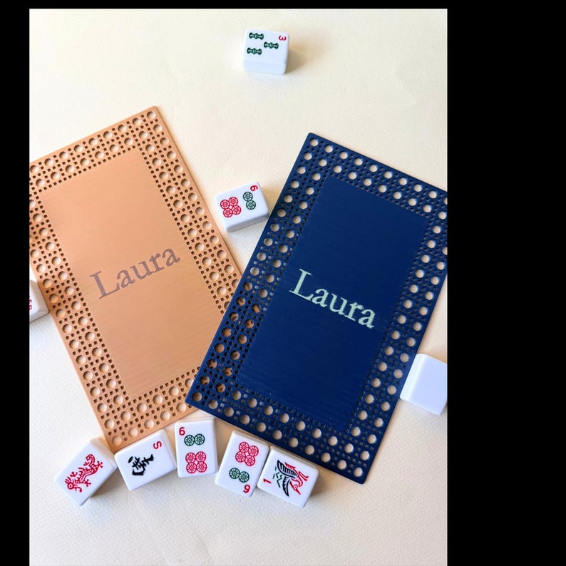Pu&ograve; includere: Due porta tessere Mahjong personalizzati, uno color pesca e uno blu navy, con il nome "Laura" stampato sopra. I porta tessere hanno un bordo decorativo e sono circondati da tessere Mahjong.