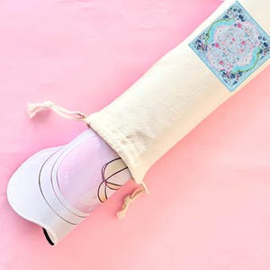May include: A rolled-up yoga mat with a floral design peeks out of a cream-colored drawstring bag. The bag has a decorative square patch with a floral pattern. The mat is pink and white with a geometric design.
