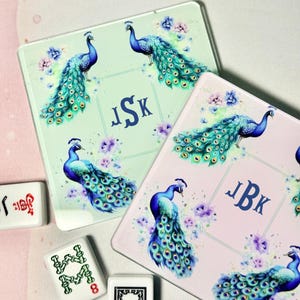 Könnte beinhalten: Zwei quadratische Untersetzer mit Pfauenmotiven. Einer ist hellgrün, der andere rosa. Jeder zeigt einen Pfau mit blauen und türkisfarbenen Federn, floralen Akzenten und einem Monogramm. Mahjong-Steine ​​sind im Vordergrund.