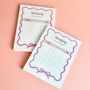May include: Two white Mahjong scoreboards with pink decorative borders and ribbon accents. Each board has a grid for tracking scores, with the words "Mahjong Score board" at the top. The background is a solid pink color.