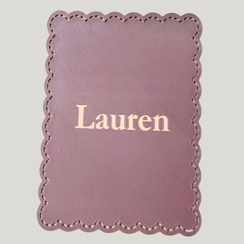 May include: A mauve rectangular coaster with a scalloped edge and the name "Lauren" printed in gold. The coaster has a stitched border and a textured surface.