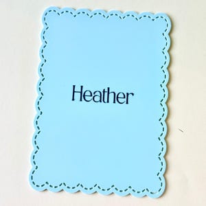 May include: A pale blue, scalloped-edge rectangle with the name "Heather" printed in black. The border has a decorative stitched design. The background is a light cream colour.