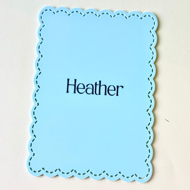 May include: A pale blue, scalloped-edge rectangle with the name "Heather" printed in black. The border has a decorative stitched design. The background is a light cream colour.