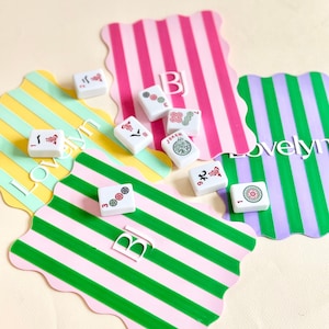 May include: A collection of white Mahjong tiles with red, green, and black symbols, scattered on colorful striped placemats. The placemats feature pink, green, yellow, and purple stripes. The word "Lovelyn" is visible on one placemat.