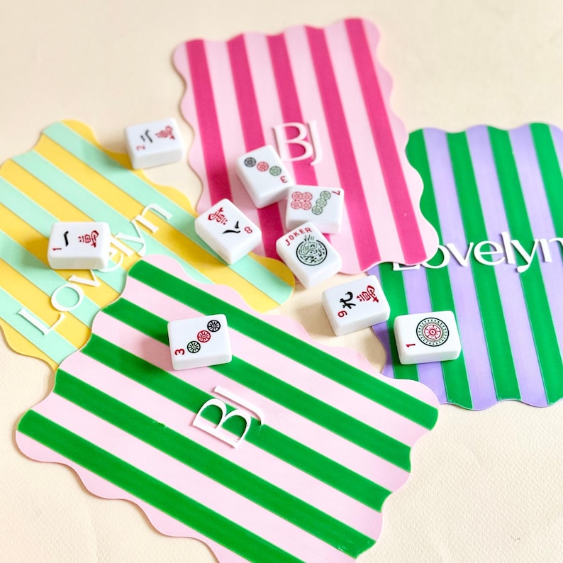 May include: A collection of white Mahjong tiles with red, green, and black symbols, scattered on colorful striped placemats. The placemats feature pink, green, yellow, and purple stripes. The word "Lovelyn" is visible on one placemat.