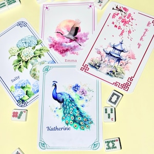 Pode incluir: Um conjunto de cartas decorativas com ilustrações em aquarela. Os desenhos incluem um pavão, flores, um grou e uma pagoda. Cada carta tem um nome. Peças de Mahjong espalhadas.