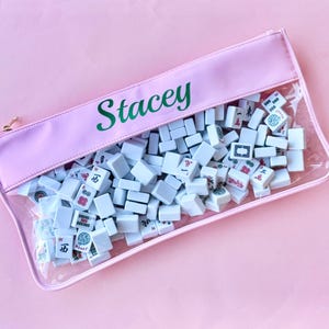 May include: A pink pouch filled with Mahjong tiles. The pouch has a clear front and the name "Stacey" in green script. The tiles are white with various symbols and characters. The pouch is zipped closed with a gold-colored zipper.