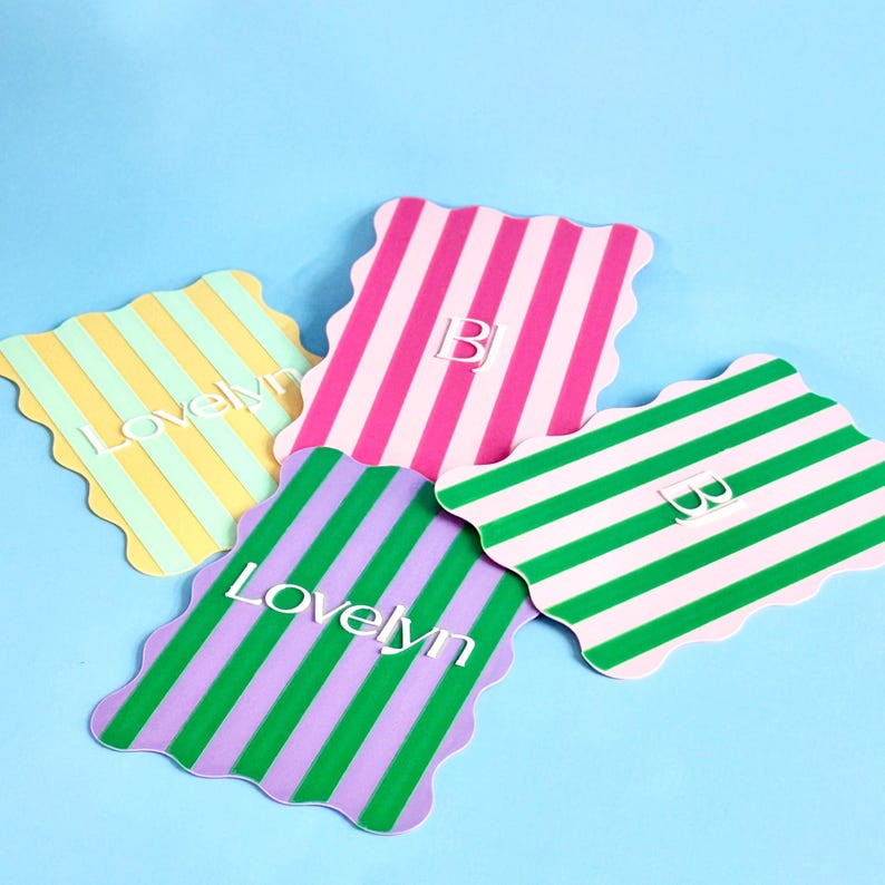 May include: Four colorful, striped coasters with scalloped edges are arranged on a blue surface. The coasters feature vertical stripes in pink, green, yellow, and purple, with white accents. Two coasters have the letter "B" and two have the word "Lovelyn".