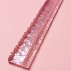 May include: A long, straight, pink acrylic hair accessory with a pearlescent, marbled texture. The piece features a decorative scalloped edge along one side and a smooth, flat surface on the other, designed for styling or holding hair in place.