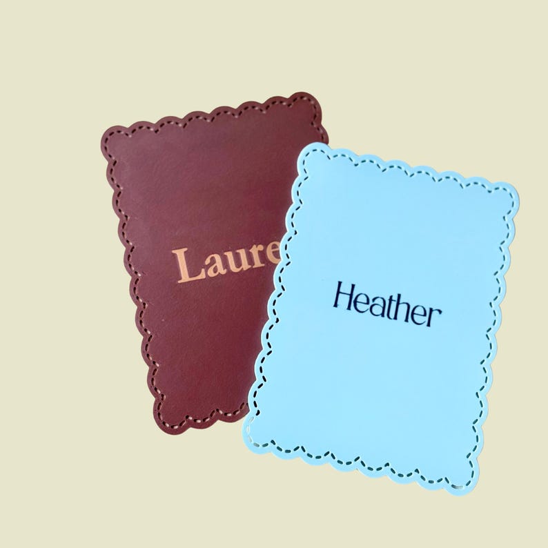 May include: Two personalised coasters. One is burgundy with the name "Laure" in gold lettering. The other is light blue with the name "Heather" in black lettering. Both coasters have a scalloped edge and a stitched border.