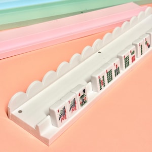 May include: A white mahjong tile rack featuring a scalloped top edge and a long horizontal channel for holding tiles. Several white mahjong tiles with red and green printed symbols and numbers are arranged in the rack, displaying various suits.