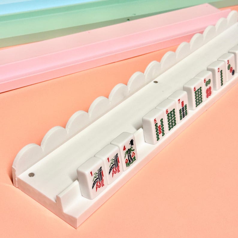 May include: A white mahjong tile rack featuring a scalloped top edge and a long horizontal channel for holding tiles. Several white mahjong tiles with red and green printed symbols and numbers are arranged in the rack, displaying various suits.